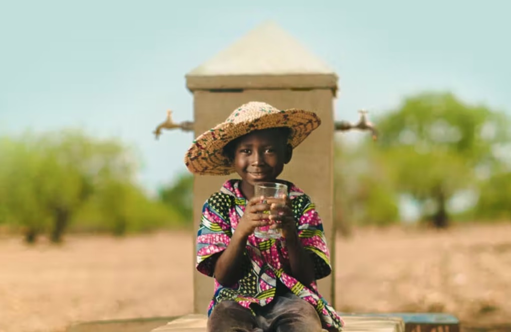 Image of a boy with a hat on, holding a glass of water and smiling