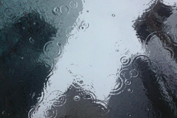 Raindrops ripple in a puddle that reflects a gray cityscape of skyscrapers.